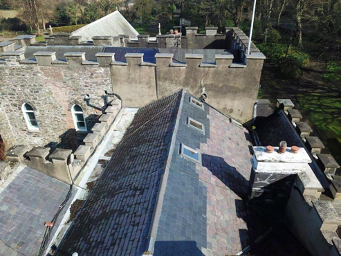 An aerial photograph showing the roof of a historic stone building with crenellated parapets, slate tiling, and skylights. The image captures chimney stacks and the surrounding rural landscape.