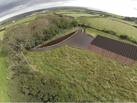 An aerial photograph showing a rural site with a large outbuilding featuring a dark roof, a curved driveway, and surrounding green fields and hedgerows.