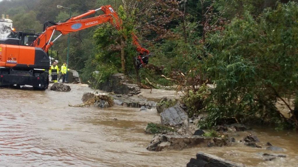 A large orange excavator is clearing vegetation and debris in a flooded, muddy area with workers observing nearby.