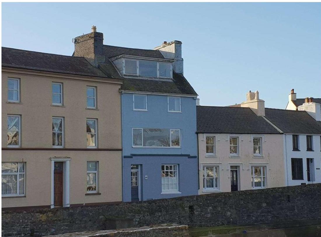 A street-level photograph showing a row of terraced houses, featuring a central blue house with a dormer window and modern windows situated behind a stone wall.