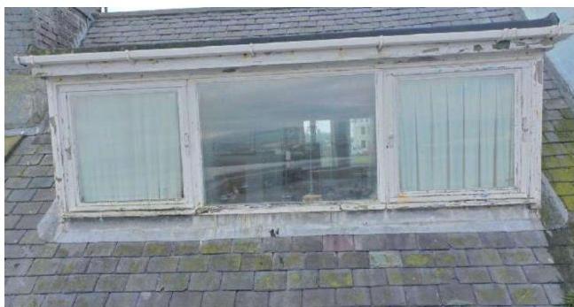 A close-up photograph of a weathered white wooden dormer window structure on a slate roof.