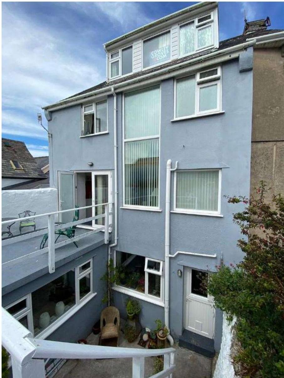 Exterior photograph of a multi-story residential building painted light blue, featuring a balcony, large windows, and a ground floor patio area.