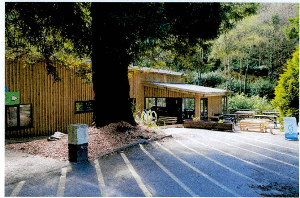 A photograph showing a single-story timber building with vertical cladding and a flat roof overhang. The structure is situated next to a paved parking area with white lines, surrounded by trees and greenery.