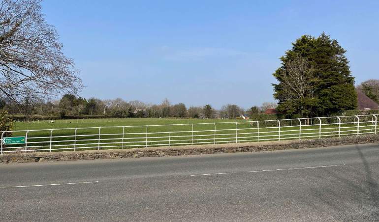 A photograph of a rural roadside scene featuring a white metal fence, a green pasture, and trees in the background.