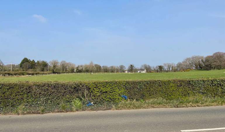 A photograph showing a rural landscape with a road in the foreground, a thick hedge, and a large green field extending to a line of trees and distant buildings.