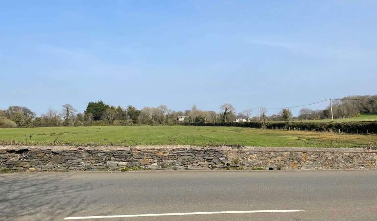 A photograph showing a rural roadside scene with a stone wall, a grassy field, and distant trees and buildings under a clear blue sky.