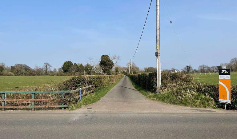 A photograph of a rural road scene showing a paved path leading into fields, flanked by hedges and a wooden fence, with a TT sign visible on the right.