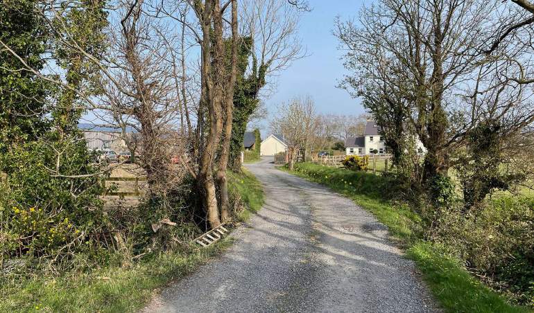 A photograph showing a gravel access road leading uphill through a rural setting with trees and buildings visible in the background.