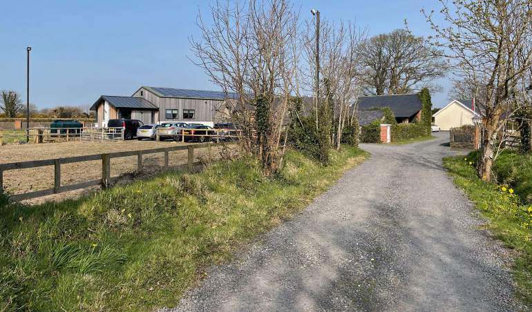 A photograph of a rural property featuring a gravel driveway and a large barn-style building with vehicles parked in front.