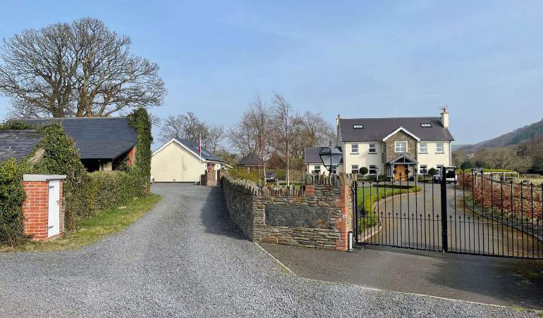A photograph showing a rural property entrance with a gravel driveway, stone wall, and black metal gate, featuring a slate-roofed building on the left and a large white detached house on the right.