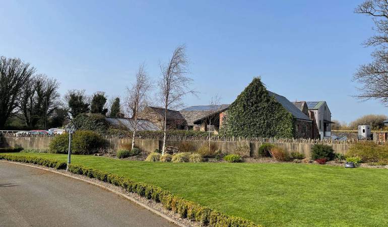 A photograph showing a rural property with stone buildings, some covered in ivy and others with solar panels, situated behind a stone wall and wooden fence.