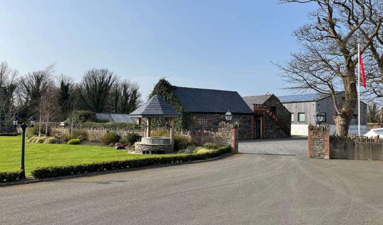A photograph of a rural property featuring a stone building, a covered well, a paved driveway, and a modern outbuilding under a clear blue sky.