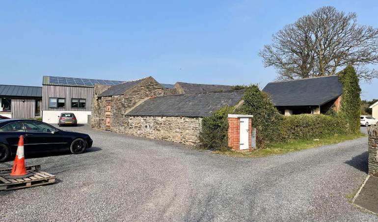 This photograph shows a rural property with a gravel driveway, a parked car, and a mix of traditional stone buildings alongside a modern structure with solar panels.