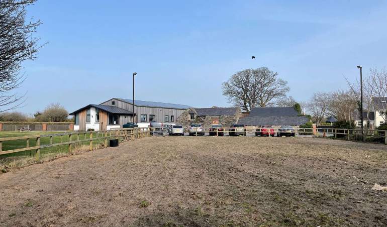A photograph of a rural site showing a modern grey building and older stone outbuildings behind a wooden fence with several parked cars.