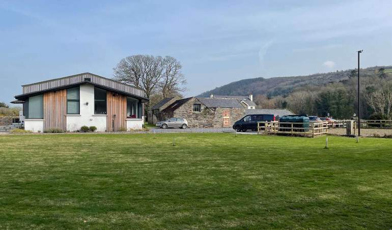 A photograph showing a modern single-story building with timber cladding adjacent to an older stone building, set within a rural landscape with a large grassy area.