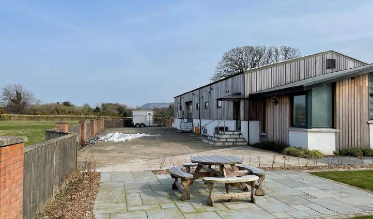A photograph showing the exterior of a long, single-story building with corrugated metal cladding and a paved yard area featuring a picnic table.