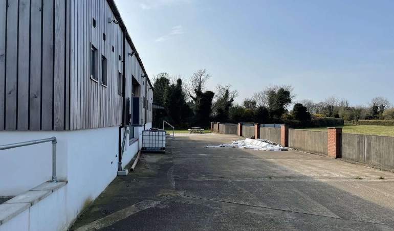 A photograph showing the side elevation of a wooden-clad workshop building with a paved yard area and boundary fencing.
