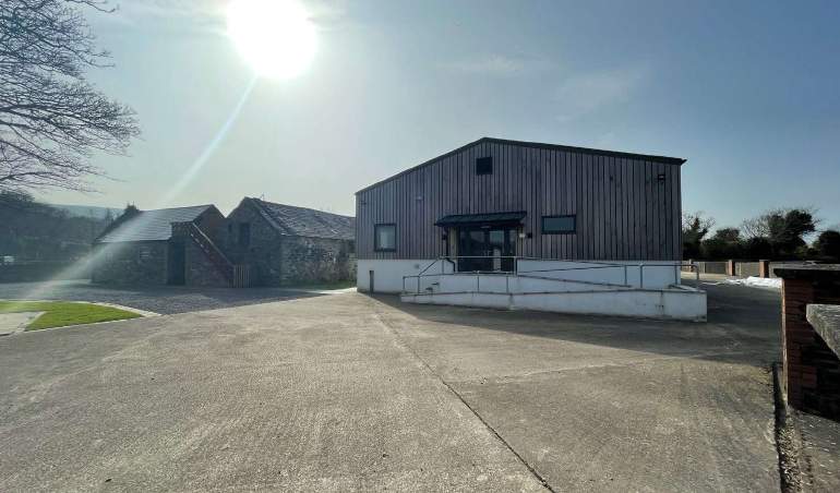 A photograph showing a modern wooden-clad building with a white base and ramp, situated next to older stone outbuildings on a rural site with a large concrete driveway.