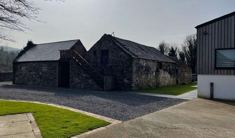 A photograph showing a stone outbuilding with a slate roof and external wooden stairs, situated next to a modern cladded building with a gravel driveway in the foreground.
