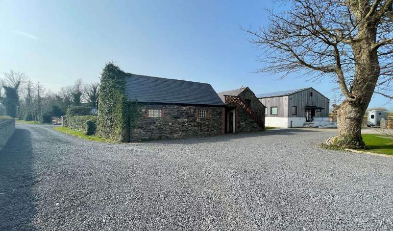A photograph of a rural property featuring a stone workshop building covered in ivy and a modern grey outbuilding, set against a large gravel driveway.