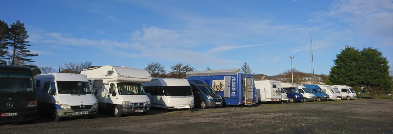A wide photograph showing a row of various motorhomes and campervans parked on a gravel surface under a blue sky.