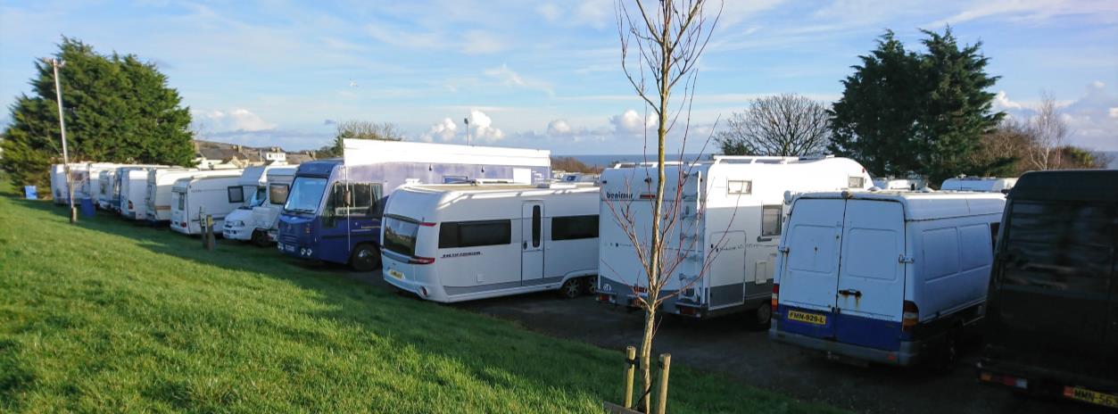 A photograph showing a row of motorhomes and vans parked on a grassy area with a coastal view in the background.