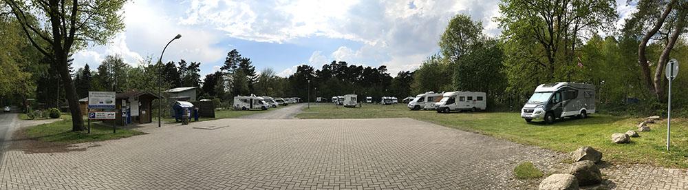 A panoramic photograph showing a paved area and adjacent grassy field where several motorhomes and campervans are parked.