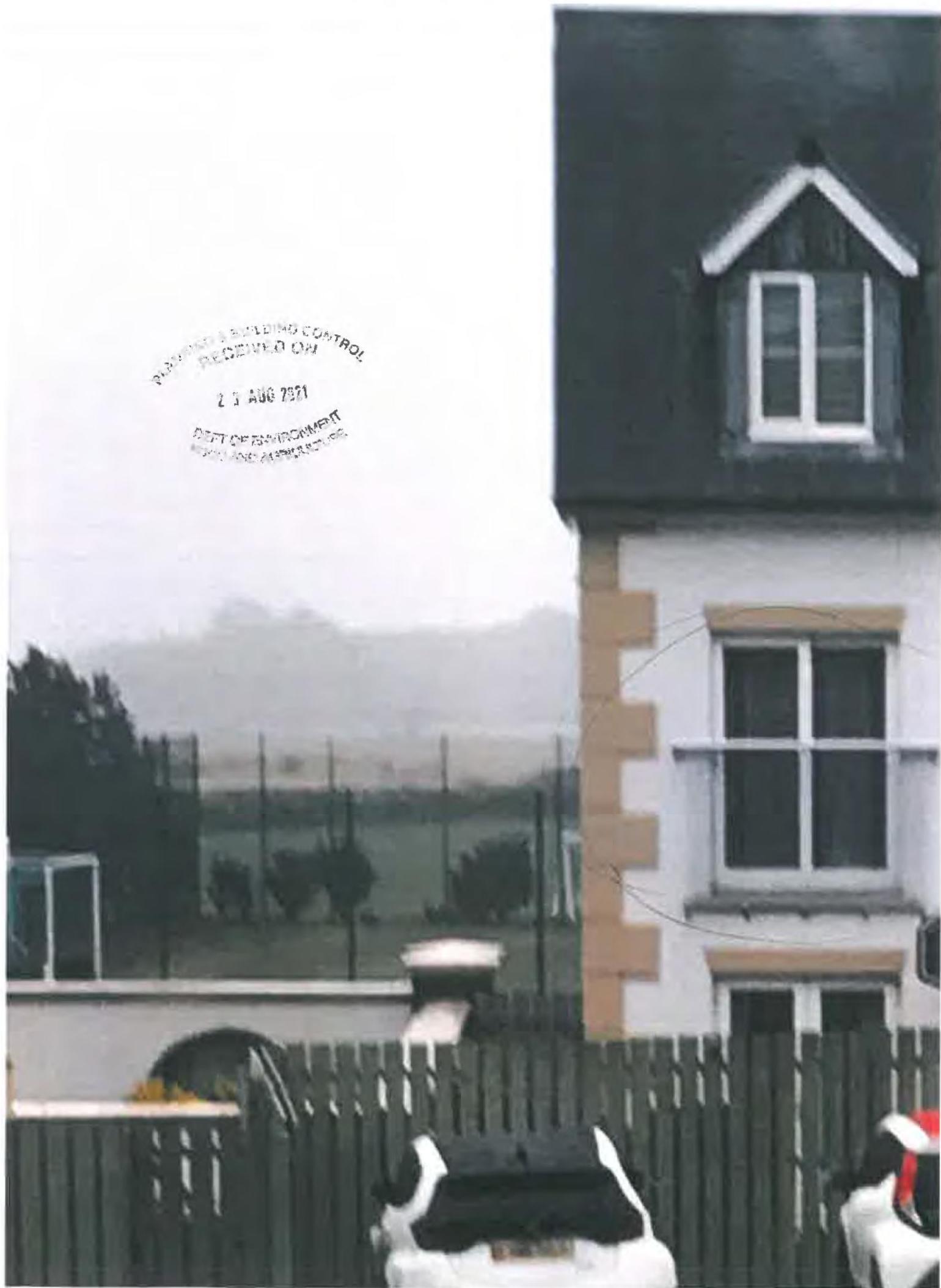 A photograph showing the side elevation of a white house with a dark roof and dormer window, viewed from behind a wooden fence with a hazy landscape in the background.