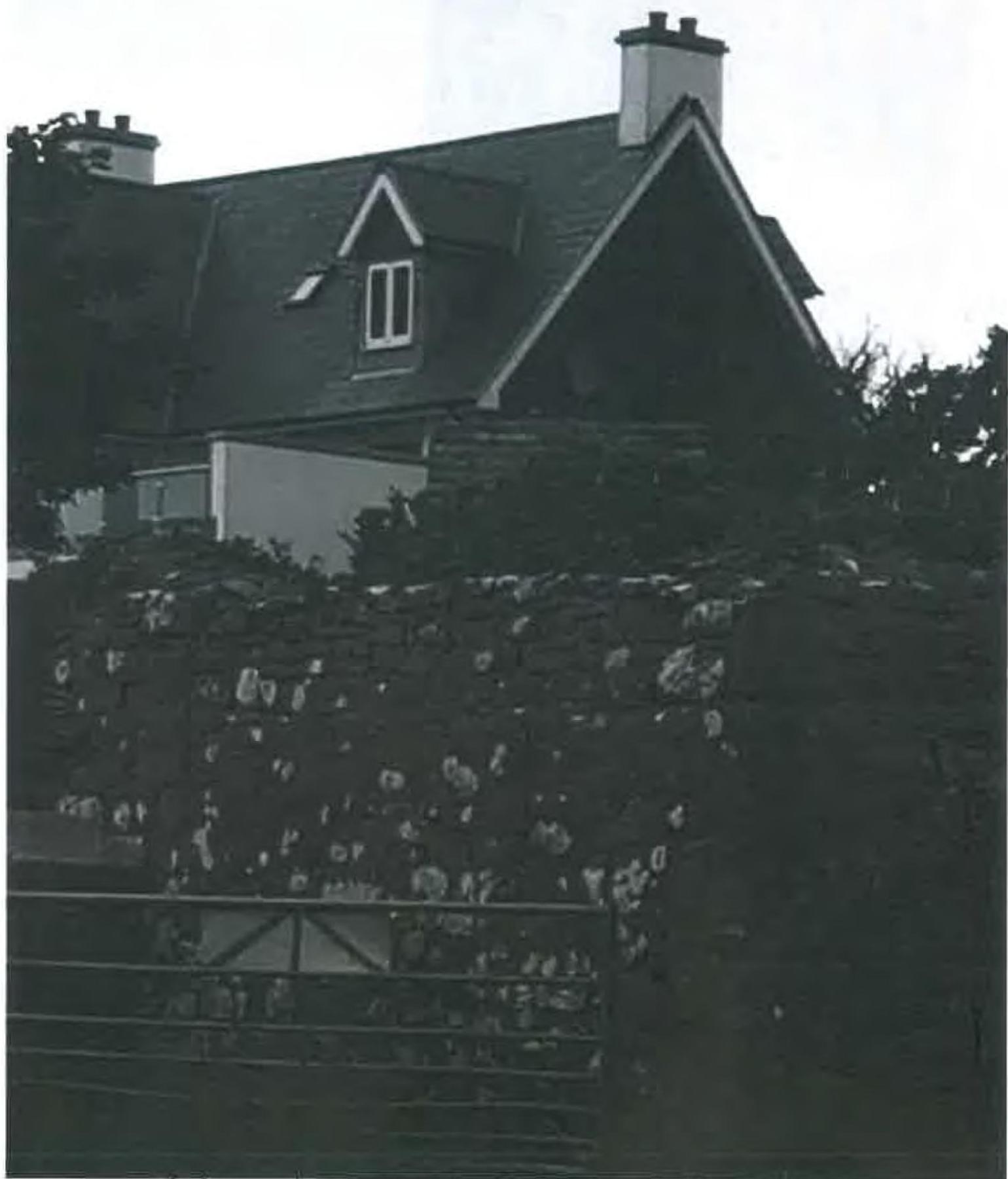 A grainy black and white photograph showing a house situated on a slope behind a large stone retaining wall, with a single-storey structure visible at the lower level.