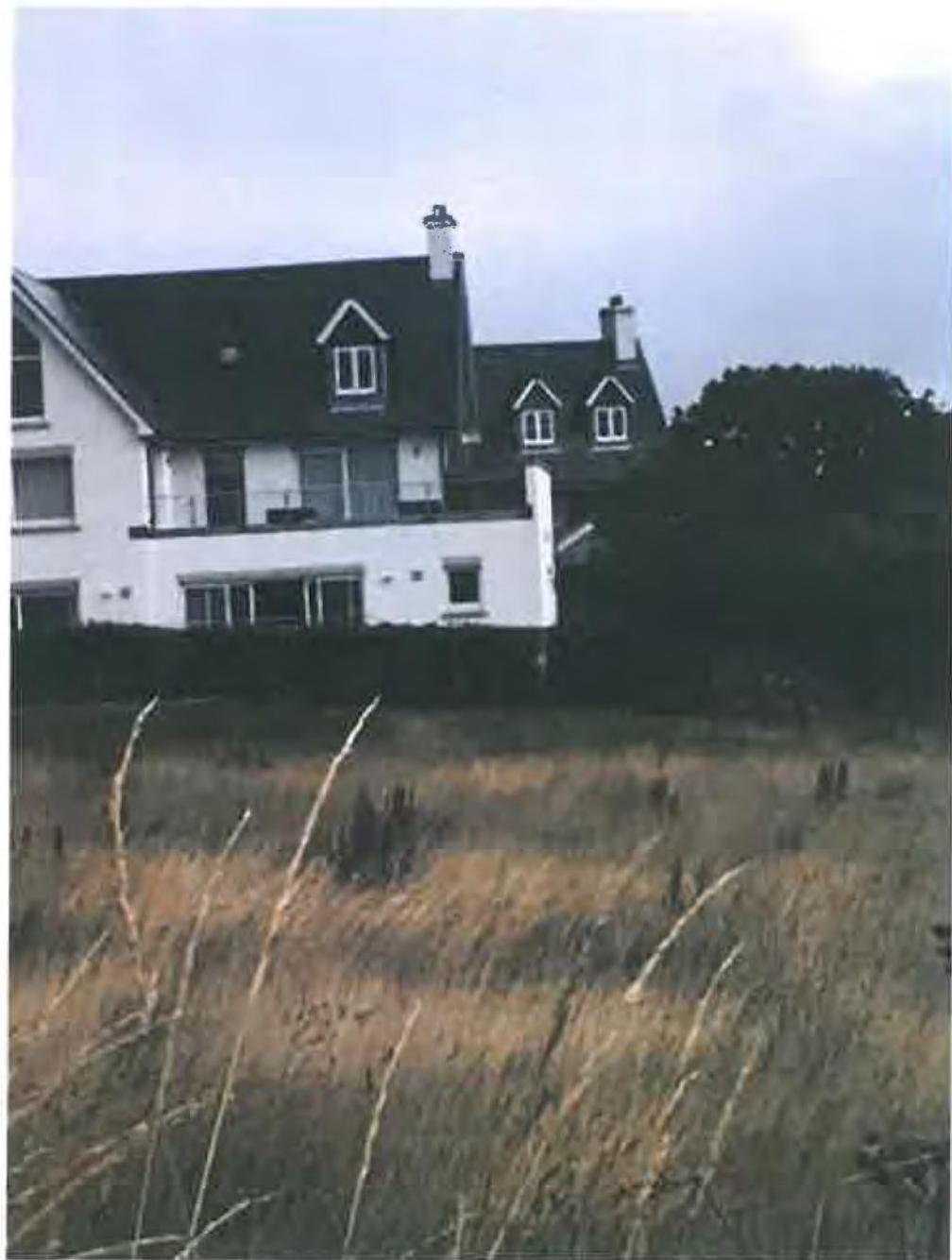 A photograph showing a white two-story house with a dark roof and dormer windows, situated behind a field of tall grass.