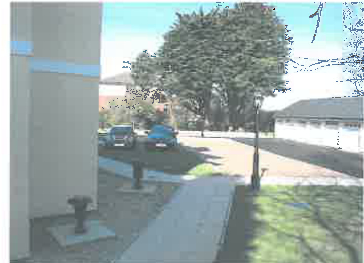 A low-resolution photograph showing a street scene with parked cars, a driveway, and buildings in the background.