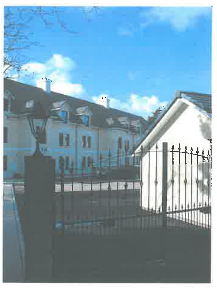 A photograph showing a white garage structure behind a black metal fence, with a larger residential building visible in the background.