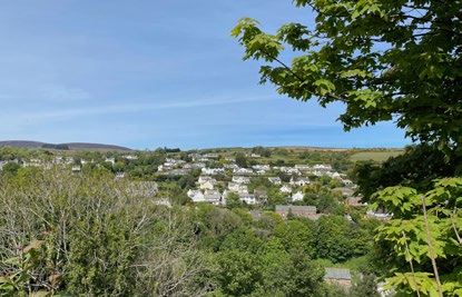 A photograph showing a scenic view of a village nestled in a green valley with houses scattered among trees, framed by foliage in the foreground.