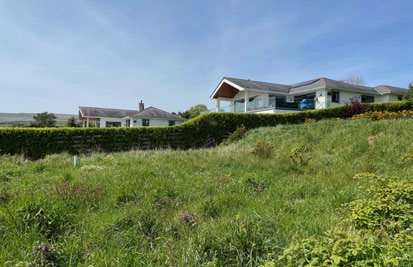 A photograph showing a grassy slope in the foreground with a hedge, behind which stand two residential properties including a modern detached house.