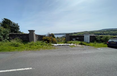 A roadside photograph showing a stone wall and gateposts with a view of the sea in the background.