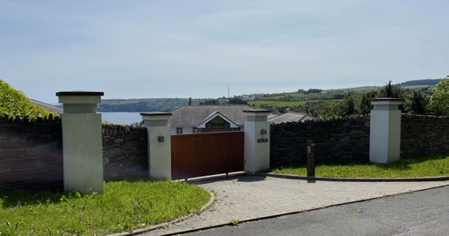 A photograph showing a property entrance featuring a stone wall, white pillars, and a wooden gate, with a house and a body of water visible in the background.