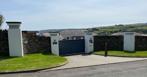 A photograph showing a stone boundary wall with white pillars and a blue gate, leading to a driveway with houses and a coastal landscape in the background.