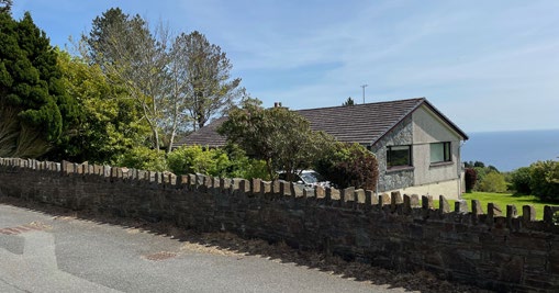 A photograph showing an existing single-story detached house behind a stone wall with a view of the sea in the background.