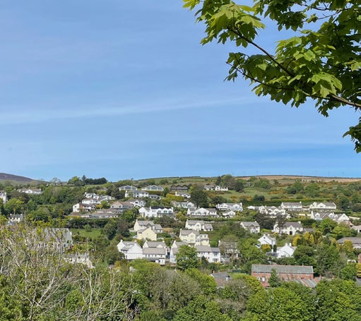 A scenic photograph showing a hillside settlement with numerous white houses nestled among green trees and vegetation under a clear blue sky.