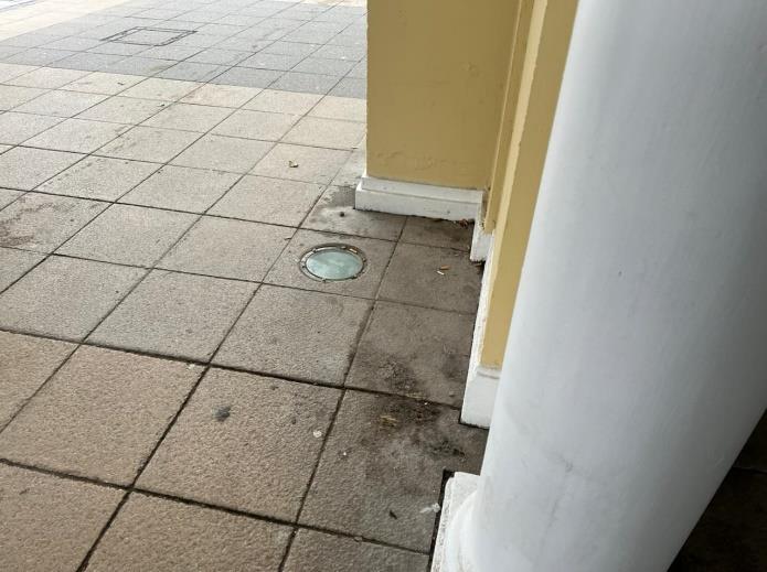 A close-up photograph of a paved sidewalk featuring square tiles, a circular ground cover, and the base of a white column next to a yellow wall.