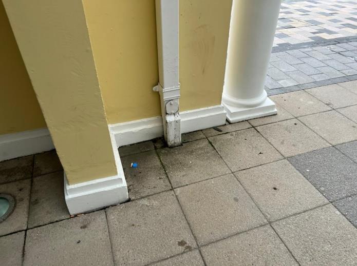 A close-up view of the base of a yellow building featuring a white downpipe, column, and paved sidewalk.