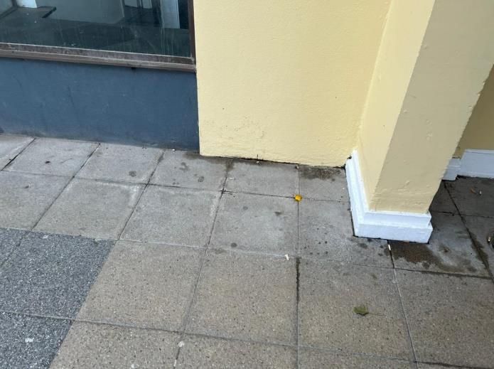 A close-up photograph showing the corner of a yellow building facade with white skirting meeting a grey paved sidewalk.
