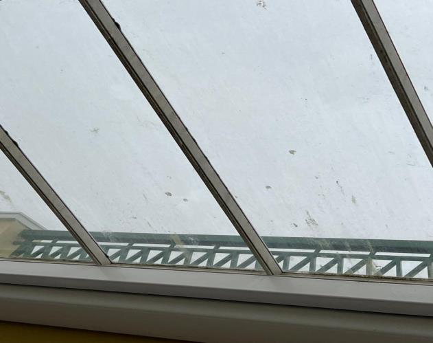 A close-up interior photograph looking up at a glass roof structure with metal framing and a green railing visible below.