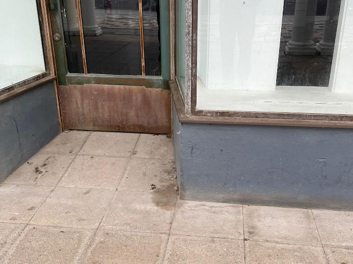 A photograph showing the ground-level exterior of a commercial property, featuring a glass door with a wooden kickplate and a large window adjacent to a paved sidewalk.