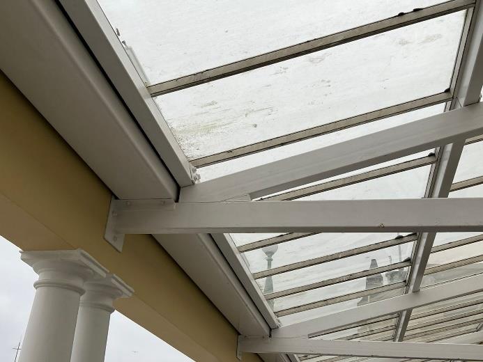 A close-up photograph looking up at a white metal framed glass roof structure, likely a conservatory or porch, attached to a yellow wall with decorative white columns.
