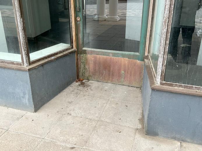 A close-up photograph of a commercial storefront entrance featuring glass windows and a door with weathered frames and a grey plinth.