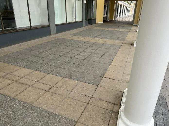 A photograph showing a paved pedestrian walkway with grey and beige square tiles. A white column stands in the foreground right, with a commercial building featuring large windows on the left.