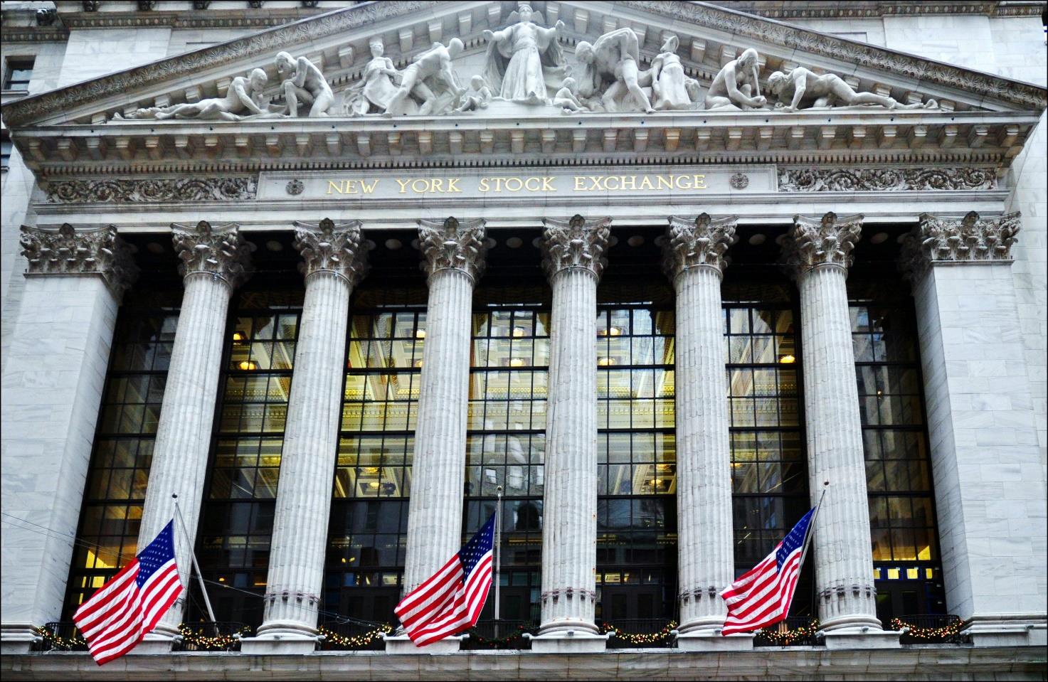 A low-angle photograph of the facade of the New York Stock Exchange, featuring large Corinthian columns, a sculpted pediment, and American flags.
