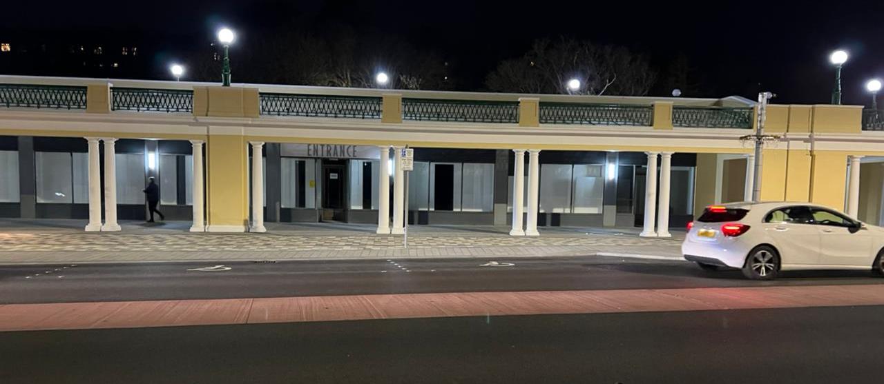 Night-time exterior photograph of a single-story commercial building featuring a colonnade and an entrance sign, with a white car parked nearby.
