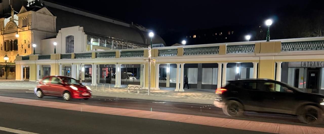 A night-time photograph of a street scene featuring a single-story commercial building with a colonnade and balcony, identified as 'Café Villa', with cars driving past on the road.